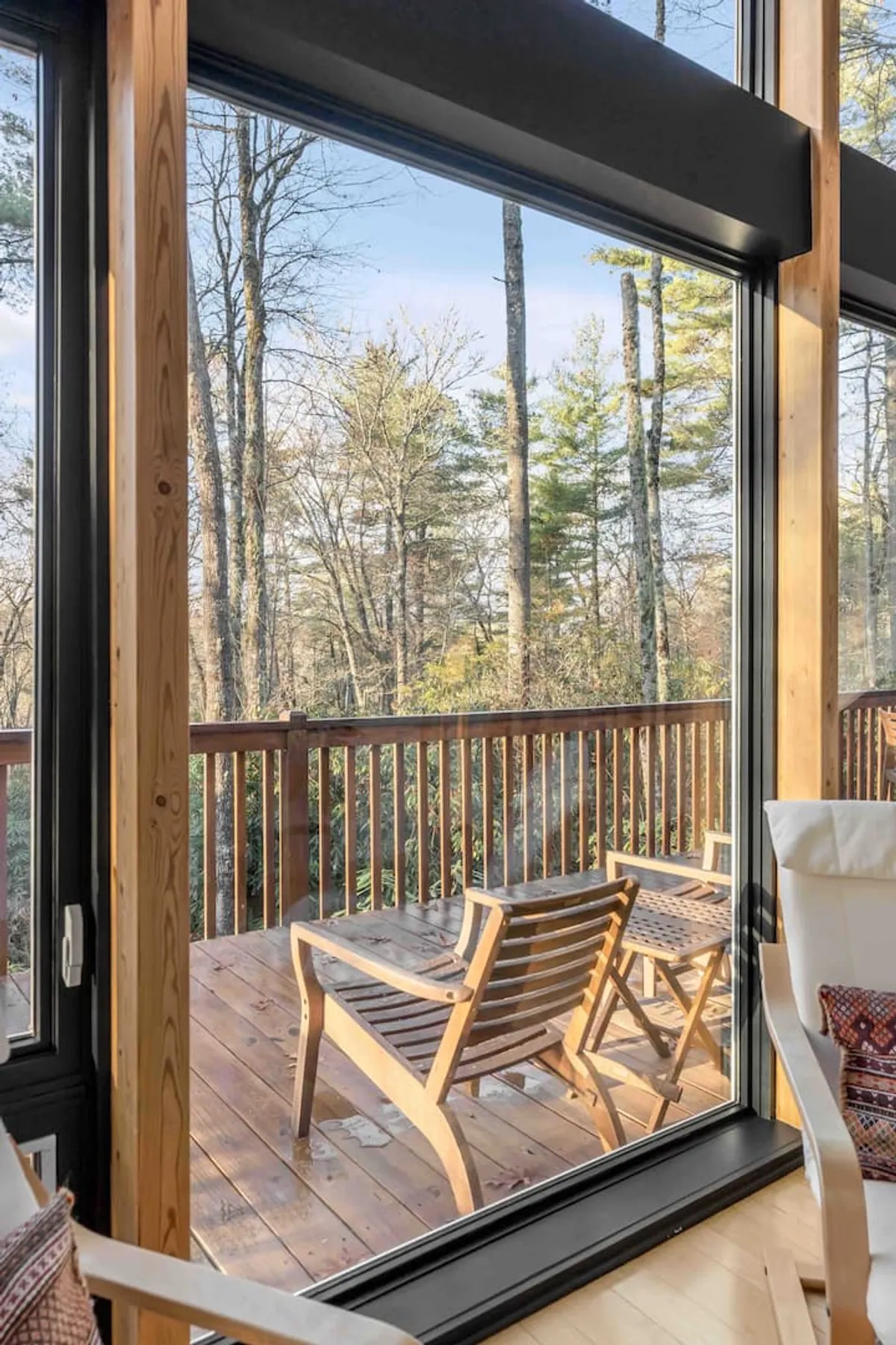 A cozy interior with large glass windows opening to a wooden deck, featuring comfortable chairs arranged for relaxation or reading. The space is filled with natural light, offering views of the outdoors.