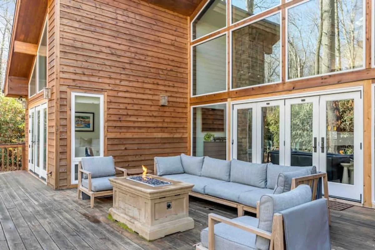 This image showcases a wooden deck with a cozy fire pit surrounded by ample outdoor seating. Large windows in the background hint at seamless transitions between indoors and outdoors, while the lush forest encircles the living area, providing privacy and natural beauty. The space is ideal for gatherings, stargazing, or enjoying a tranquil morning coffee.