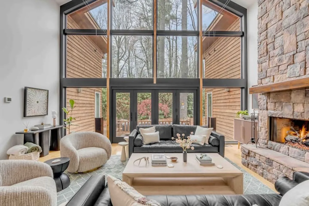 The hero image showcases a cozy yet modern living area featuring a dramatic stone fireplace, sleek black leather sofa, a light wood coffee table, and impressive floor-to-ceiling windows that bathe the room in natural light and frame views of the surrounding forest.
