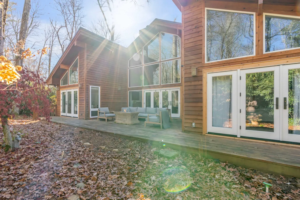 This tranquil scene features a spacious wooden deck with comfortable outdoor seating surrounded by towering trees. Large windows on the house reinforce the indoor-outdoor connection, and the scene suggests both morning coffee and evening gatherings are equally inviting here.
