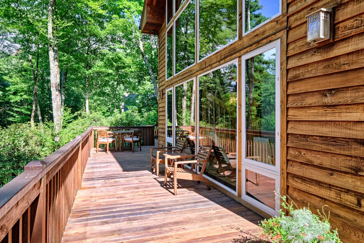 An inviting view of the home’s outdoor wooden deck with large windows, comfortable outdoor furniture, and a backdrop of surrounding trees. The tranquil setting suggests a seamless connection to nature, with opportunities for morning coffee or evening relaxation in peaceful seclusion.