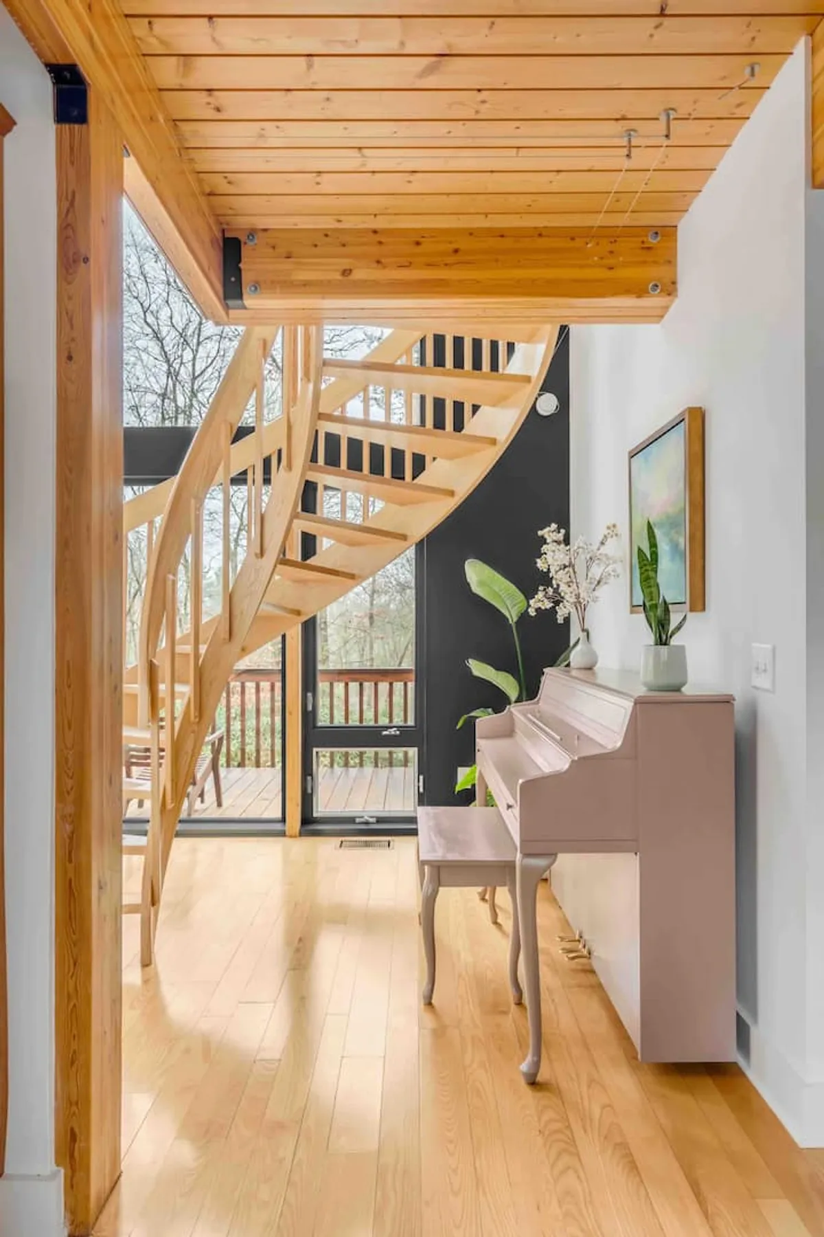 An inviting space featuring a spiral staircase, a pink piano, and large windows that let in plenty of sunlight. The room exudes creativity and overlooks greenery outside, enhancing the inspirational feel of the loft area.