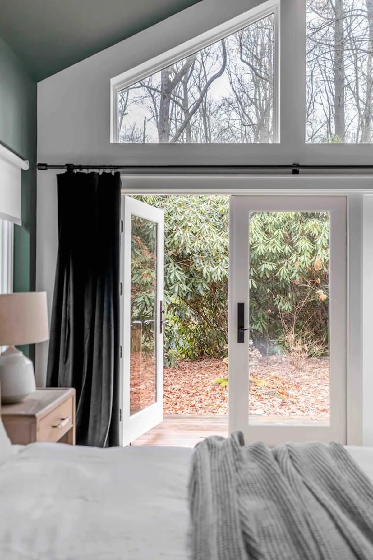 A guest bedroom with open French doors leading to an outdoor space, a bed adorned with a textured blanket, and a simple nightstand with a lamp. Natural light floods the room, creating an airy, restful ambiance.
