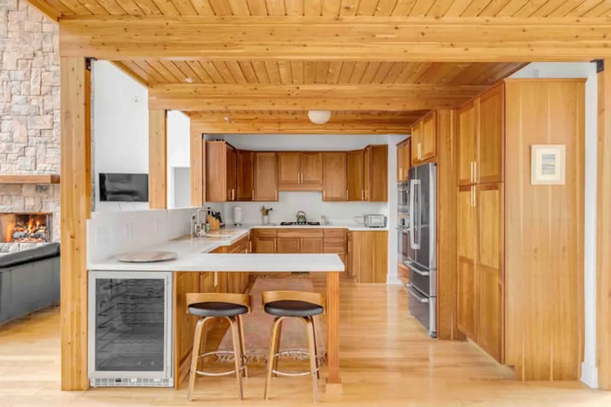A chef-ready kitchen opens into the living space, featuring sleek wooden cabinetry, a central island with seating for casual chats, and a stone fireplace in the background. The kitchen is set up for both culinary adventures and conversation, with ample natural light filtering in.