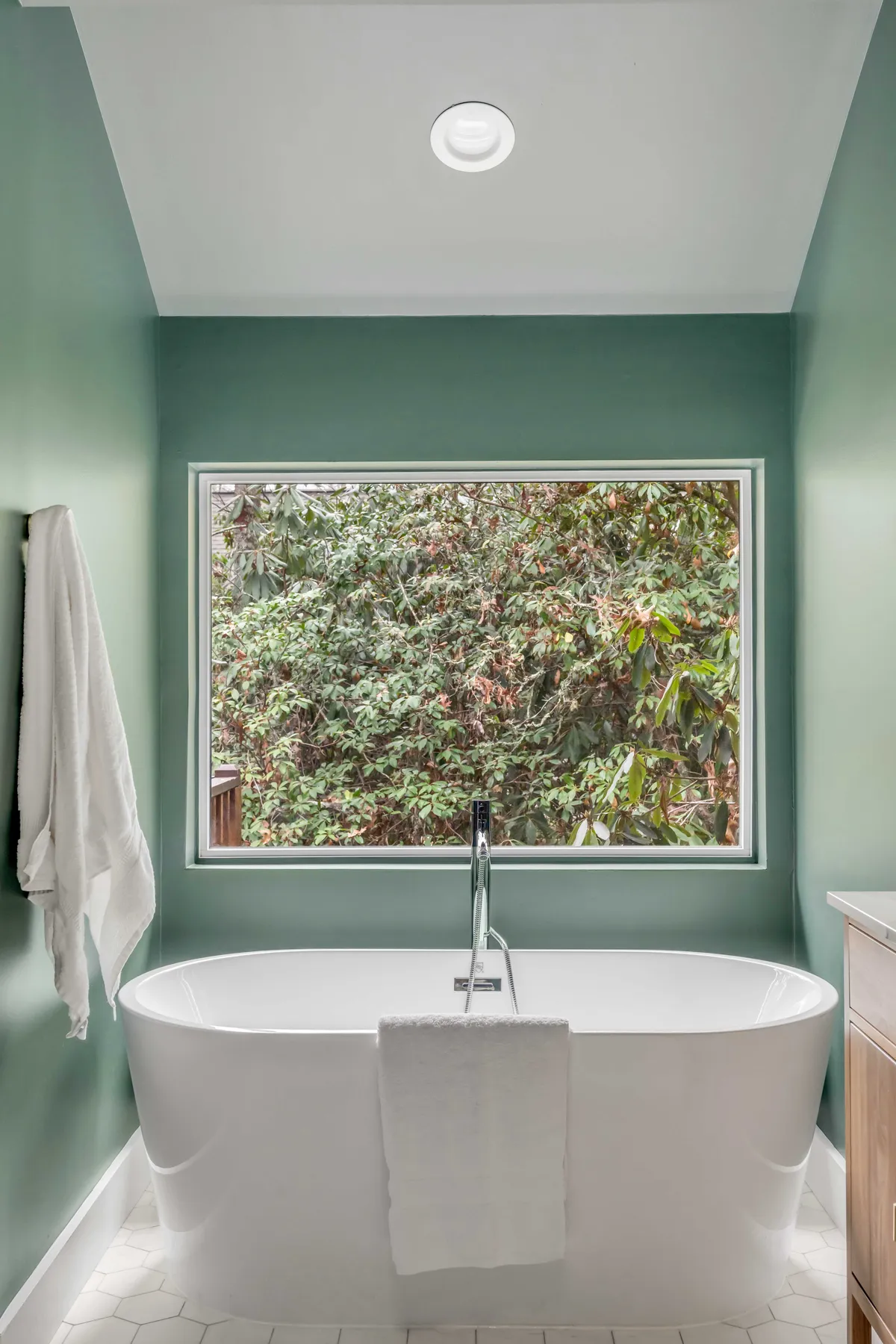 A spa-inspired bathroom with a freestanding bathtub positioned by a large window, green walls, and tranquil natural views. The scene radiates relaxation and the luxury of a private spa.