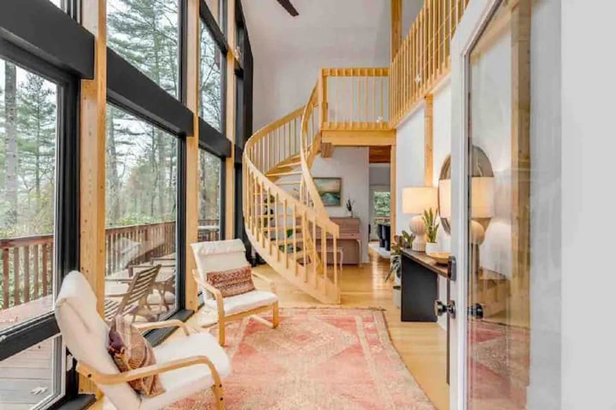 This image captures a spiral staircase leading up to a sunlit loft with large windows, a comfortable seating area, and a decorative rug. The view includes natural light streaming in, enhancing the tranquil and creative atmosphere of the space.