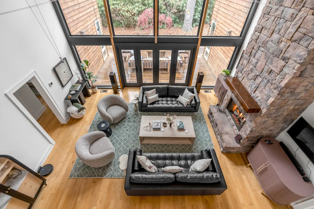 A warm living area featuring a large stone fireplace, cozy seating with plush cushions, a stylish coffee table, and expansive floor-to-ceiling windows that frame lush forest views. Natural light pours into the open-plan space, highlighting the inviting and social atmosphere.