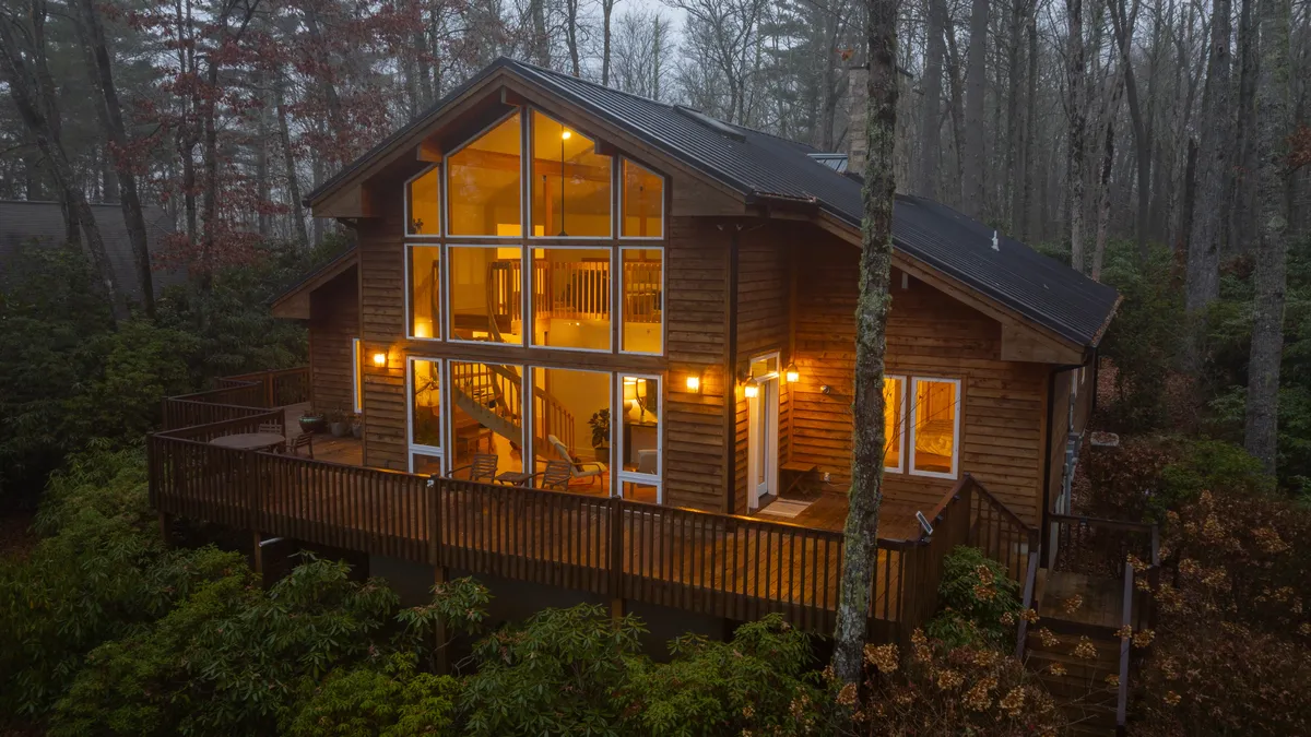 This image showcases the chalet's impressive glass windows and wooden deck, surrounded by lush trees. The outdoor fire pit area suggests a perfect spot for evening gatherings, all set within a tranquil, forested landscape. The interior staircase is also visible, blending indoor comfort with outdoor access.