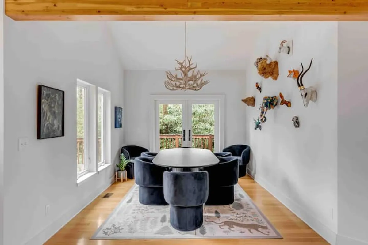 A contemporary dining area with a round table surrounded by unique, bold chairs and striking wall art. Large windows bathe the space in natural light, offering a glimpse of the forest outside—an ideal setting for memorable group feasts or intimate celebrations.