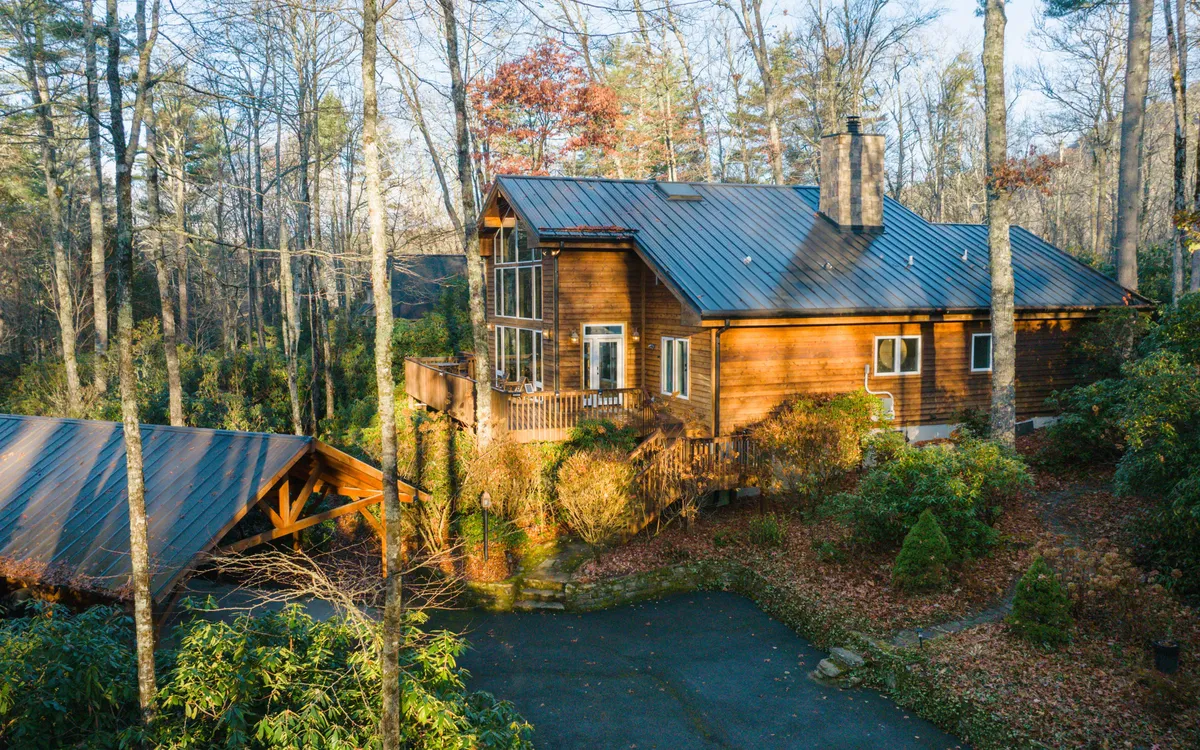 A wooden house is nestled among tall trees, with a spacious deck area in the foreground. The scene is set at sunset, creating a warm, inviting glow across the patio. There are comfortable seating areas visible, ideal for gathering and relaxing outdoors as evening falls.