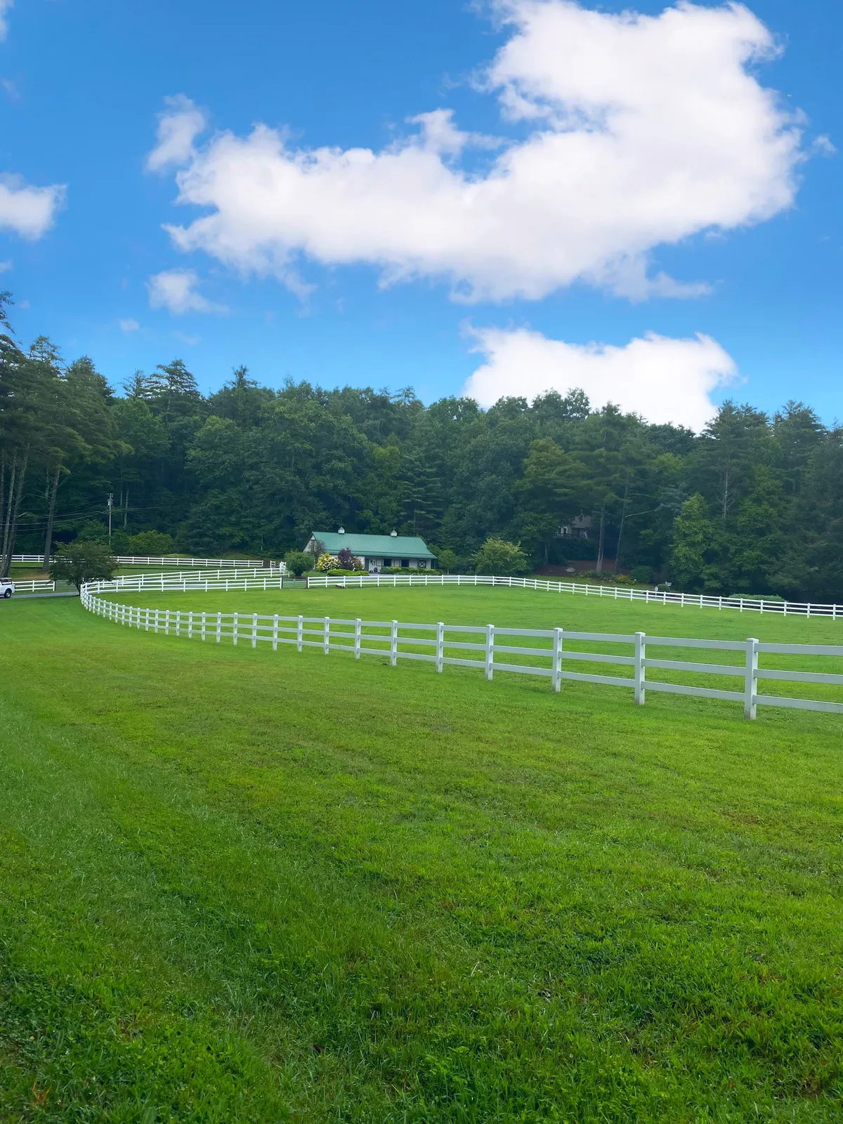 A panoramic view of green pastures bordered by a white fence, with a charming cottage-style house and a forested background. The open lawn provides an idyllic setting for outdoor gatherings or quiet stargazing under the evening sky.