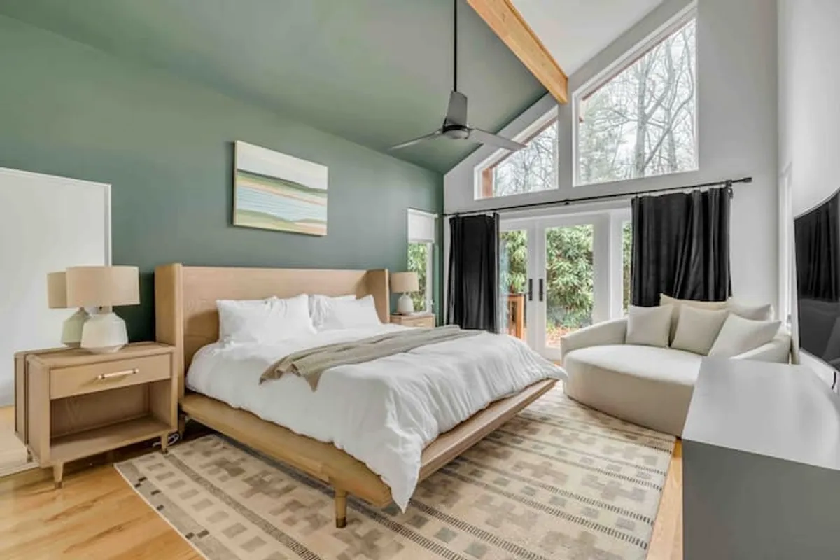 A calm and inviting bedroom with a large bed, expansive windows allowing plenty of natural light, a cozy sitting area, and thoughtfully chosen artwork. The serene palette and soft textures underscore a sense of relaxation and privacy.